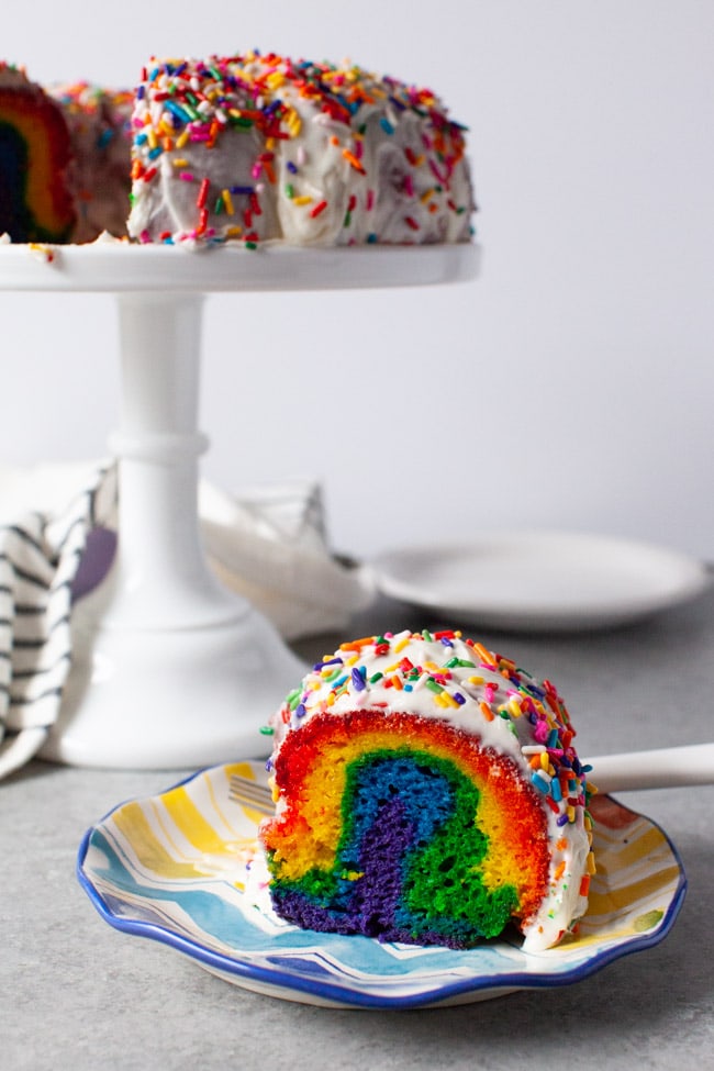 Rainbow Bundt Cake from thelittlekitchen.net Slice of rainbow cake on a plate next to tray of cake