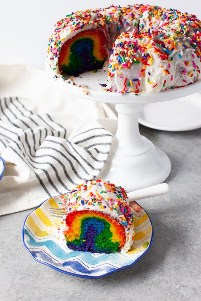 Rainbow Bundt Cake from thelittlekitchen.net Slice of rainbow cake on a plate next to tray of cake