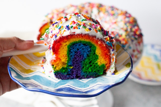 Rainbow Bundt Cake from thelittlekitchen.net Slice of rainbow cake on a plate with the full cake in the background